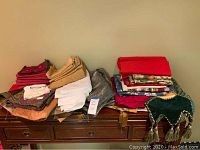 Photo showing multiple folded table linens including various colored and patterned cloths, towels, and runners on a wooden surface.