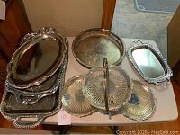 Overview of silver plated 3 tiered folding tidbit server and several silver plated serving trays on table