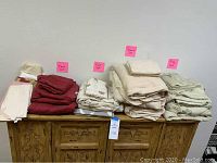 Four folded bed sheet sets arranged from left to right: Twin (burgundy), Full (off-white with emblem), Queen (beige), King (light cream/tan)