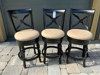Set of three black wood swivel bar stools with beige cushioned seats, decorative X-back with central floral motif, turned legs with circular footrest, arranged on outdoor stone surface.