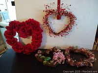 Photo showing four heart-shaped wreath decorations on a black table and hanging on a wall; includes a large red rose wreath, grapevine wreath with berries, and two smaller floral wreaths.