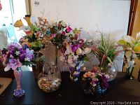 Photo of multiple faux floral arrangements in various glass vases on a black surface with a white backdrop.