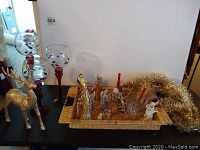 Photo showing three red stemmed wine glass style candleholders with holly pattern, gold reindeer figurine, basket containing figurines and gold tinsel garland on a surface against a wall.
