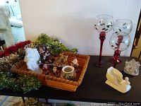 Photo showing decorative basket with multiple small Santa figurines and Christmas ornaments, white ceramic sleigh figurine, and three tall glass candleholders with red stems.