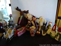 Photo showing a brown plush moose, various Santa figurines, snowmen, decorative sleighs, nutcracker, and holiday-themed vases and containers arranged on a surface.