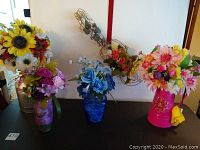 Front view of five faux floral arrangements in decorative containers on a black surface against white background.