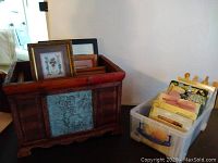 Wooden box filled with assorted picture frames and framed art including floral prints, alongside a plastic container holding additional framed items and collectibles.
