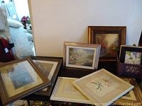Multiple framed prints and frames arranged on a table by a wall, showing different sizes and styles including floral and scenic images.