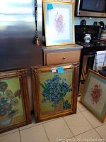 Four floral prints arranged near kitchen area; two framed prints with ornate gold-colored frames and two unframed pastel floral prints on cardboard.