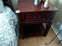 Front view of one nightstand showing drawer with metal handle and bottom slatted shelf, dark wood finish, placed next to bed with patterned bedding.