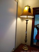 Full view of one gold leaf effect floor lamp lit, showing the base, pole and shade with tassel detail.