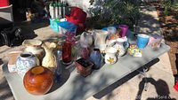 Table with a variety of ceramic and glass flower pots and jugs arranged outdoors.