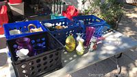 Overall view of multiple blue crates filled with assorted flower pots and holders, including ceramic, glass, and tin, arranged on a table outdoors