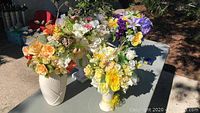 Four artificial flower arrangements displayed on a table outdoors, featuring yellow, purple, white, and orange flowers in white ceramic and green glass vases.