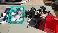 Photo showing green basket with assorted light bulbs including globe and spiral CFL types, and multiple electrical cords and chargers laid out on table.