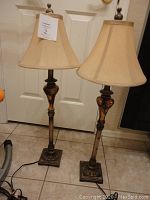 Pair of tall slender lamps shown on tile floor in front of white door, bronze textured bases and gold silk lampshades visible
