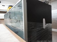 Side angle view of the tall charcoal black cabinet wrapped for protection, showing solid side panel and part of glass etched doors with aluminum frame.