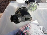 Two black Rain Drip Water Timers placed on a white surface next to the product manual, clear front covers reveal dials and connections visible.