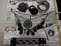Photo showing an assortment of gunmetal and black jewelry including necklaces, bracelets, and earrings arranged on a silver textured background.