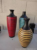 Photo of three tall decorative vases in turquoise, red, and yellow-gold with black horizontal stripes and black necks and bases, arranged outdoors on a flat surface against a brick wall.