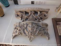 Front view of one decorative corbel showing detailed carved scroll and floral designs, approximately 16 inches wide, beige weathered finish.