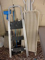 Photo showing aluminum 3-step ladder with black steps, and standard striped ironing board next to it.