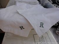 Close-up of two white linen napkins embroidered with the letter 'R'. One napkin shows a slightest discoloration or stain, the other appears clean. Both display dark green embroidery.