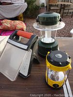 Three small camping lanterns displayed on a table: green fluorescent lantern, yellow LED lantern, and a smaller LED lantern.