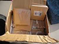 Open cardboard box filled with multiple packs of square unfinished pressed wood coasters, mostly unopened in plastic wrap.