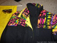 Ski jacket showing vibrant multicolor pattern on black with yellow lining and sunglasses beside yellow ski pants