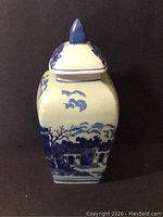 Front view of the urn showing a house with archways and clouds above in blue on white ceramic.