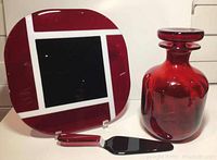 Ruby red glass decanter, cake plate with black and white geometric design, and matching glass cake cutter arranged together for display.