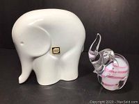 Both elephant sculptures shown side by side on black background. Larger white ceramic elephant has original Peter Muller Sgrafo Atelier label sticker. Smaller blown glass elephant with pink swirl pattern is in front right.