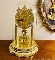 Front view of gold-tone anniversary clock with clear glass dome, showing face with Roman numerals and decorated center.