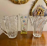 Front view photo showing three items: cut glass pitcher with handle and flower etching, colored geometric patterned vase, clear swirl design vase