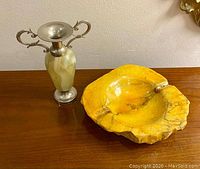 Photo of entire lot showing small onyx vase with silver-tone handles and base beside a yellow alabaster ashtray on wood surface.
