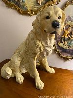 Front and side view of the light beige dog figurine showing naturalistic fur texture and sitting pose on a wood surface with decorative paintings in the background.