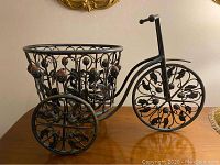 Side angle showing metal tricycle flower pot stand with decorative floral design on wheels and basket