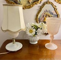 Photo showing the lamp, faux flower vase, and pink goblet on wooden surface, with ornate wall art in background