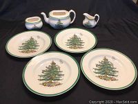 Teapot, cream jug, and sugar bowl with matching Christmas Tree pattern and green trim, shown alongside the four dinner plates on black fabric background.