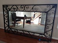 Front view of large rectangular wall mirror with ornate iron scrollwork frame showing clear reflective glass and surrounding hardwood floor and walls.