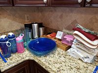 Photo showing water bottles, drinking glasses, blue plastic plates, dish towels, plastic containers, wooden flatware organizer, and kitchen tools including spiralizer and measuring cups on granite countertop.