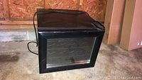Front view of black Haier wine cooler with glass door, placed on concrete floor.