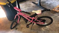 Side angle showing the full pink TREK bike with flat front tire, white and pink padded saddle, and black pedals under indoor lighting.