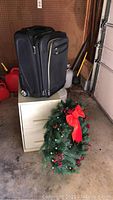 Photo of a small black Samsonite suitcase on top of a small cream-colored metal file cabinet. Artificial Christmas wreath rests in front of the cabinet; background shows a garage or storage area with gas cans and propane tanks.