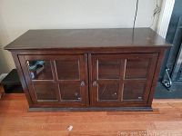 Full front view of a dark wood rectangular media cabinet with double front tempered glass doors showcasing a divided square pane design, two round knobs, and some surface scratches and dust.
