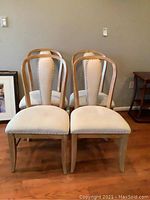 Four matching wood framed side chairs with tan upholstered seats and backs with nail trim shown arranged in two front and two back positions on hardwood floor.