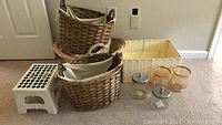 Woven baskets, white plastic step stool, two metal bathroom canisters with lids, two glass vases, and a pale yellow woven bin visible on carpeted floor.