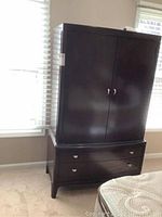 Full view of dark wood Thomasville armoire with two doors and two drawers below, situated near windows.