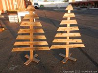 Two wooden Christmas trees standing outdoors on pavement under natural light, side by side showing overall appearance and size